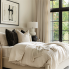 Modern bedroom with beige curtains, abstract wall art, and neutral bedding, featuring large windows.