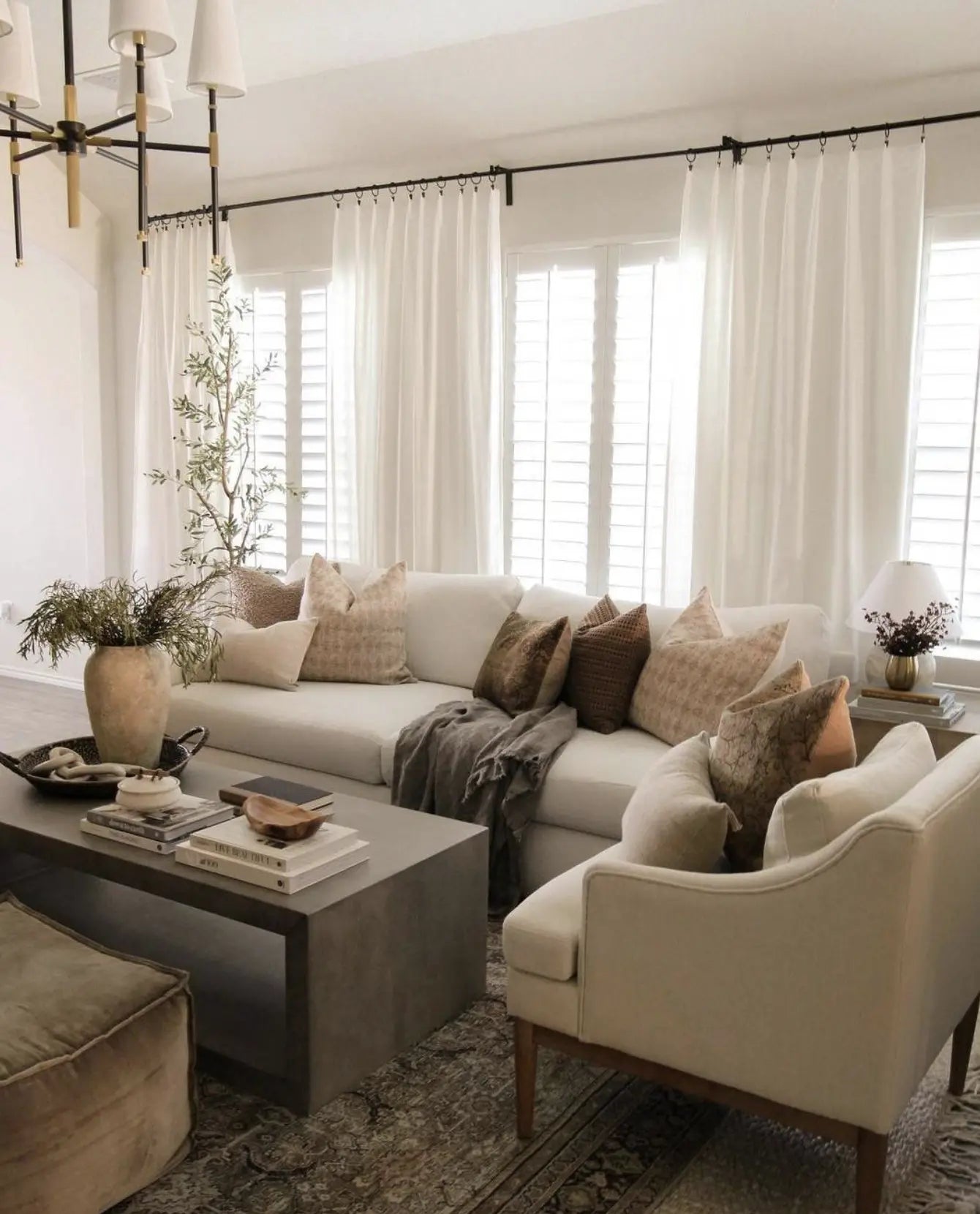 A modern farmhouse living room features premium white linen-cotton blend grommet curtains, allowing natural light to flood the space. The neutral palette complements the sofa, armchair, coffee table, rug, wooden accents, window shutter decorations, and modern chandelier, creating a cozy, inviting atmosphere.