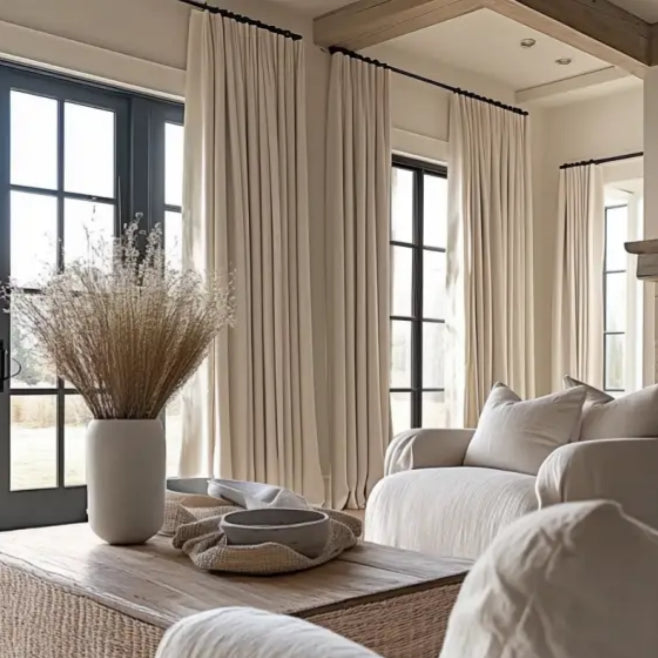 Modern living room with floor-to-ceiling cream drapes, black window frames, neutral furniture, and dried floral decor for a cozy ambiance.