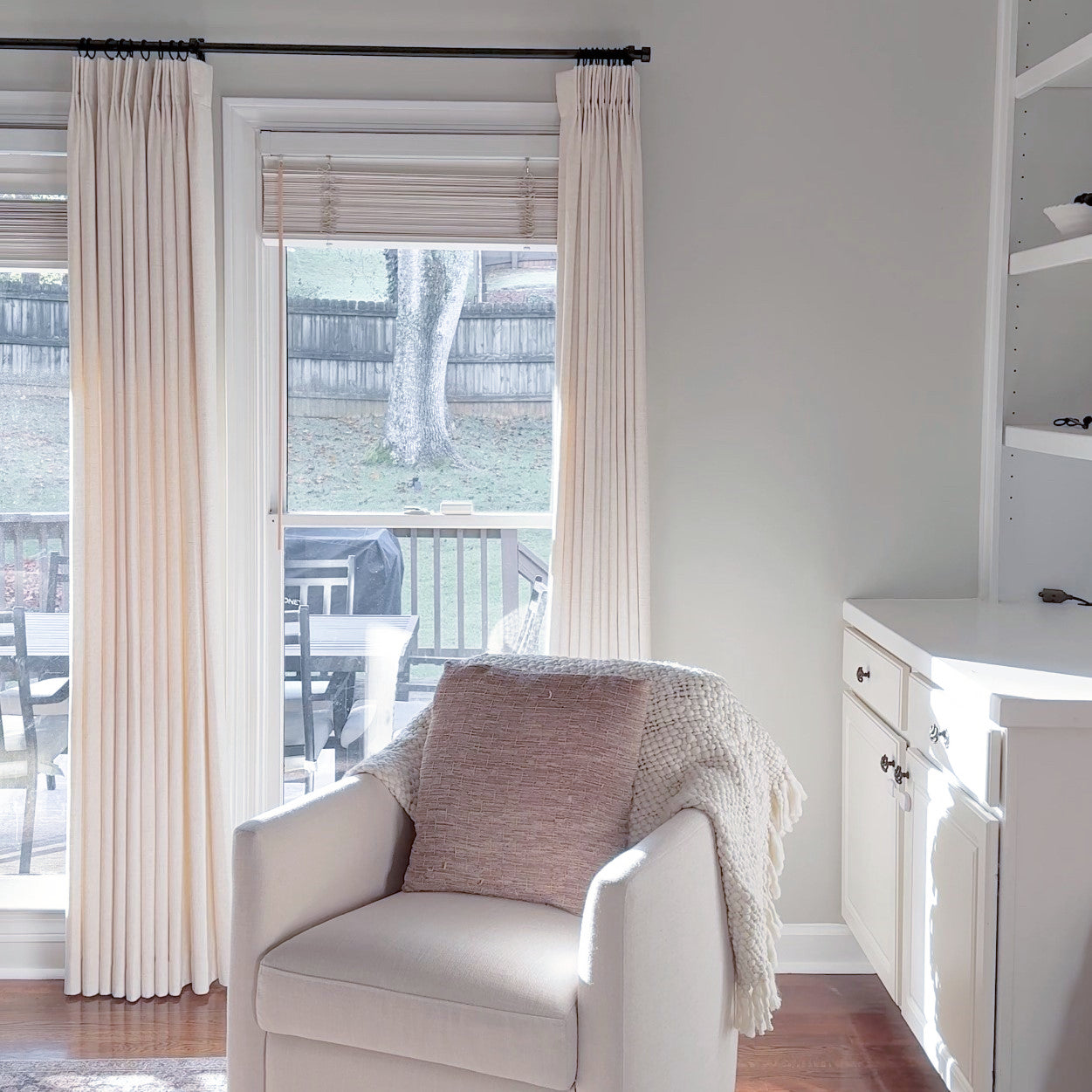 Cozy reading nook with cream armchair, textured throw, and neutral drapes framing a glass door, offering a view of a serene backyard.